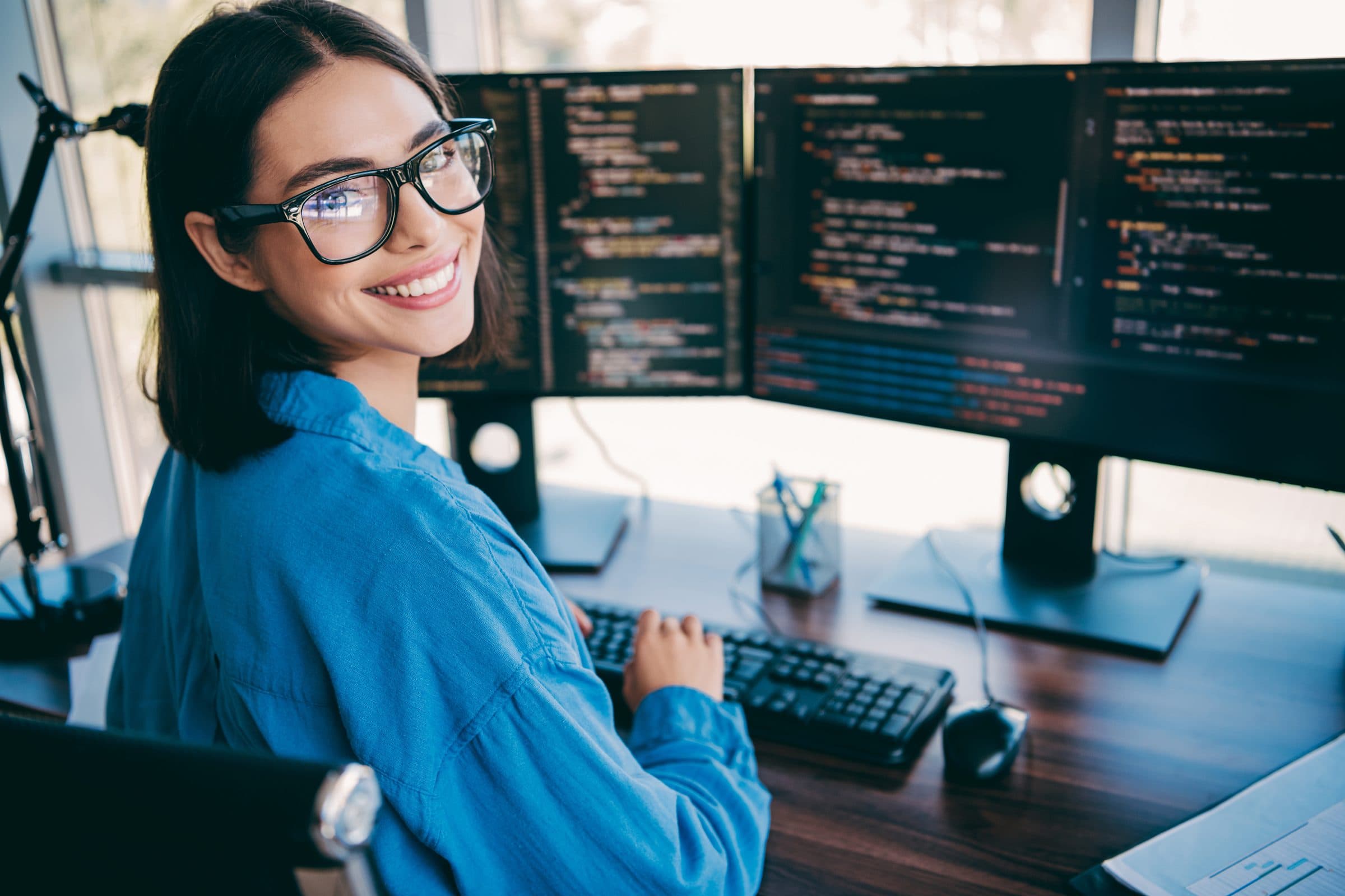 A software engineer at her desk, turning from three monitors of code to smile at the camera