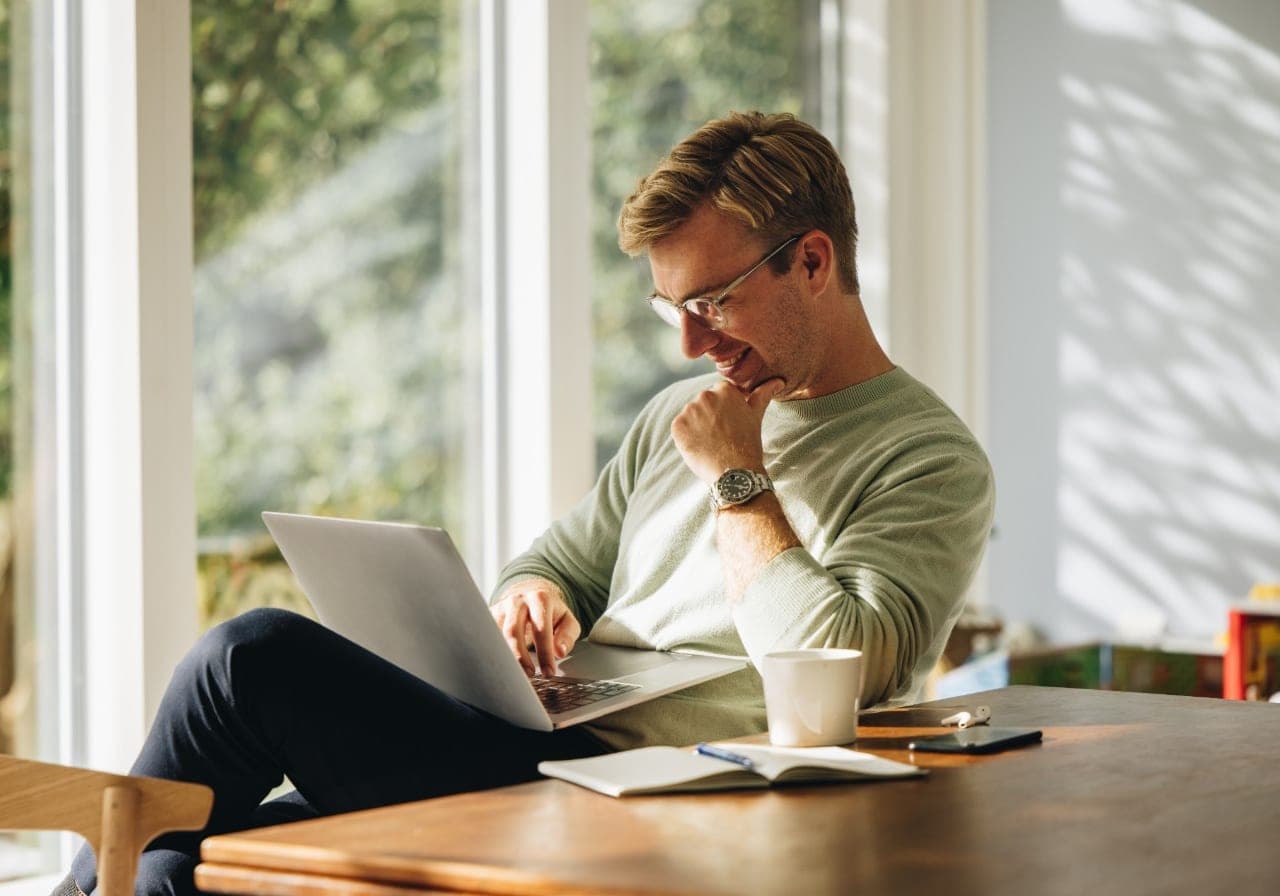 A man working on a laptop at a table by a sunlit window