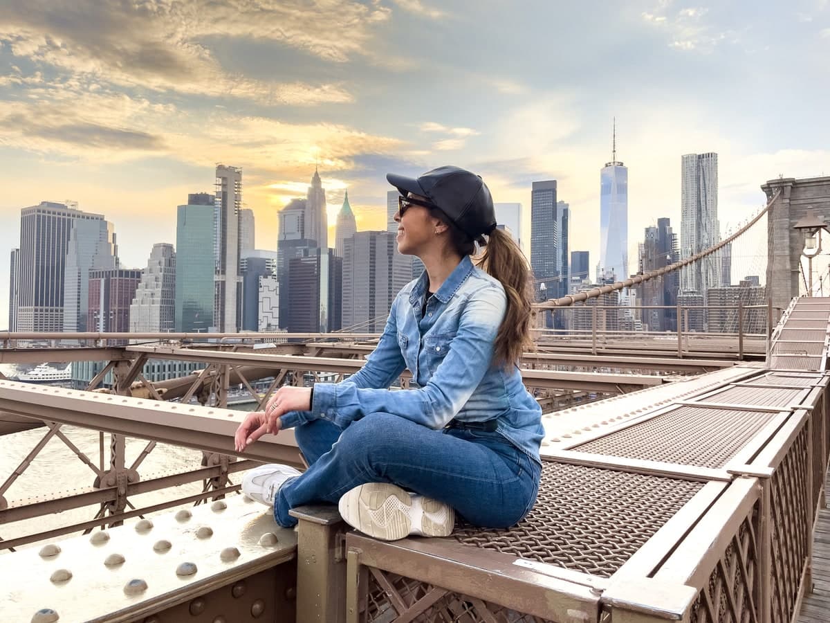 A woman sitting on the Brooklyn Bridge at sunset, the Manhattan skyline behind her